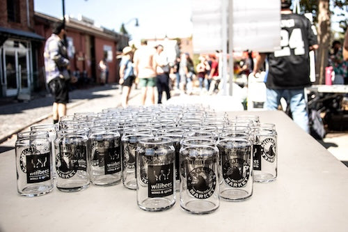 Row of mason jars beer glasses with sponsor labels from Willibees and Petaluma Market on a table outdoors, people in the background.