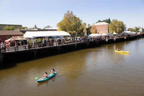 Two people in a kayak on the Petaluma river with a dock and the beer fest ents in the background.