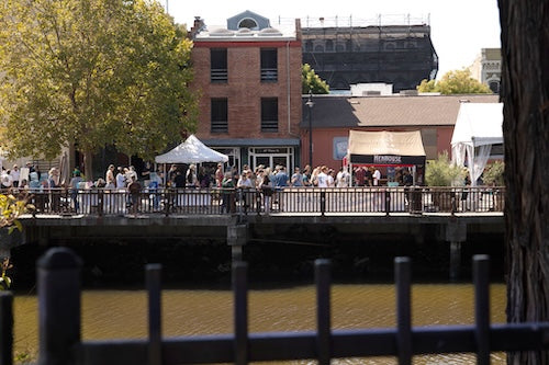 Photo of the Beer Fest from the other side of the river.  River in the foreground, tents and people in the mid-ground, Petaluma buildings in the background.