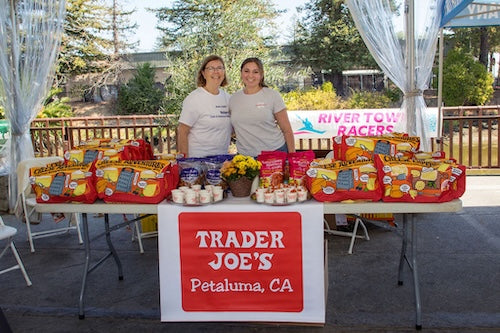 Two women standing behind a table in the Trader Joe's tent with Trader Joe's goodie bags for designated drivers on the table.