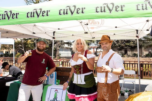 Two people posing in authentic German clothing under a 'cuVer' branded tent at an outdoor event.  Another male in regular clothes toasting for the photographr.
