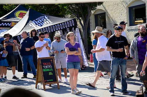 Group of people standing outdoors near beer tents with a food stand with a sandwich board pointing towards a food tent.