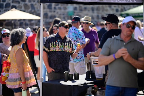 People at an outdoor event with a man in a black hawaiian shirt with beer bottles across his chest standing behind a table with bottles.