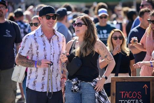A couple standing in a crowd holding beer glasses with a chalkboard sign advertising tacos and tortas.