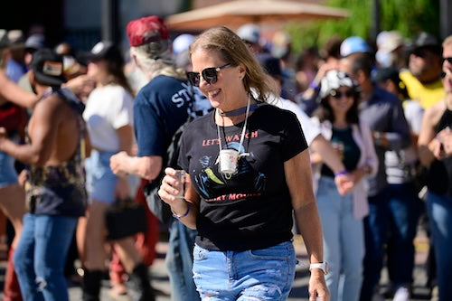 Happy woman in a crowd wearing sunglasses and a black t-shirt with the beer lanyard holding her glass around her neck.