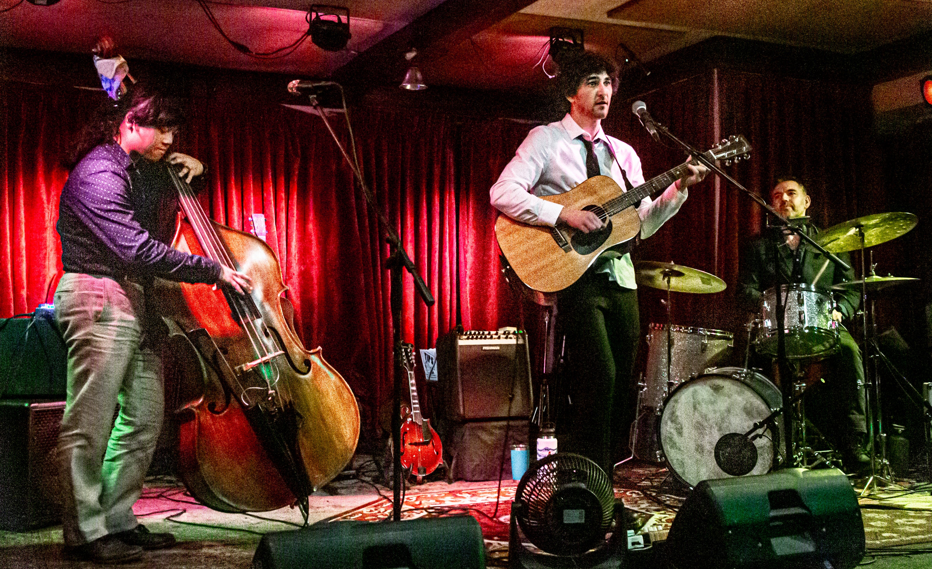 A live band performing on stage in a dimly lit venue, featuring a double bass player, a guitar player in a white shirt, and a drummer with red curtains in the background.