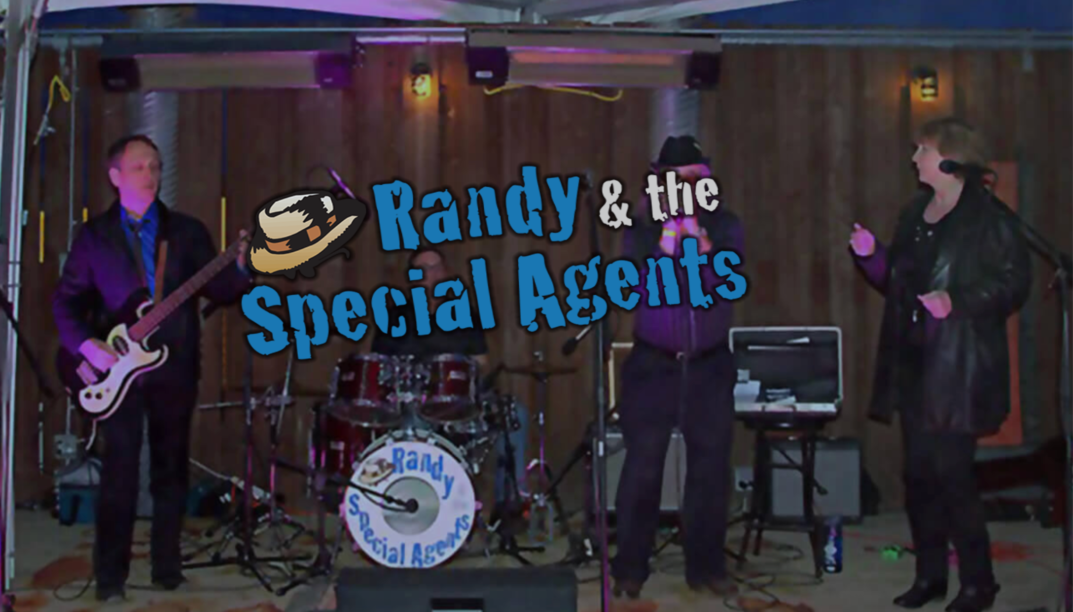 Three people performing live music on a stage under a canopy, with a large blue text overlay reading 'Randy & the Special Agents' and a cowboy hat graphic. The band members are dressed in dark clothing, with instruments including a bass guitar, drums, and a keyboard visible.